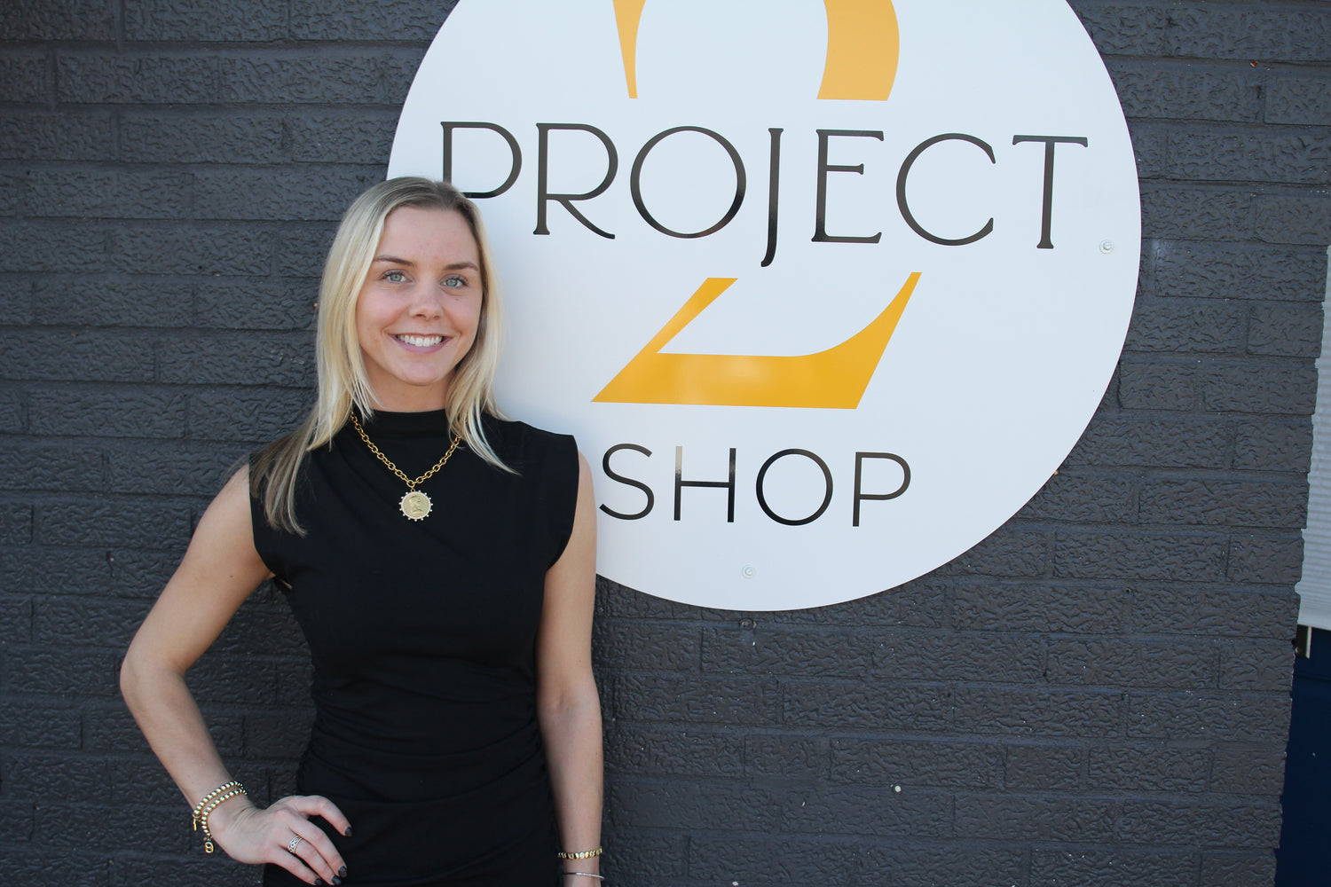 Woman standing in front of a 'Project 2 Shop' sign on a brick wall.