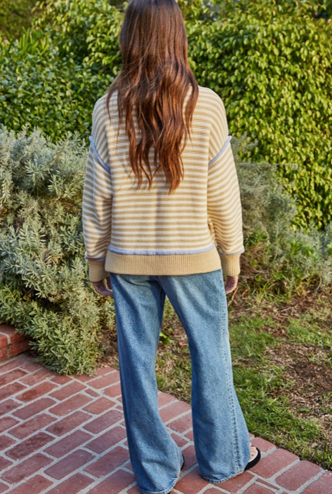 Person wearing a striped sweater and jeans standing on a brick path with greenery in the background